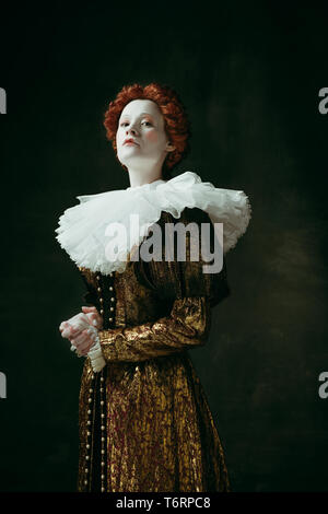 I make decisions. Medieval redhead young woman in golden vintage clothing as a duchess standing crossing hands on dark green background. Concept of comparison of eras, modernity and renaissance. Stock Photo