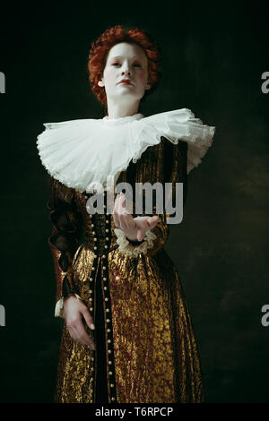 Give it to me. Medieval redhead young woman in golden vintage clothing as a duchess standing pointing up on dark green background. Concept of comparison of eras, modernity and renaissance. Stock Photo