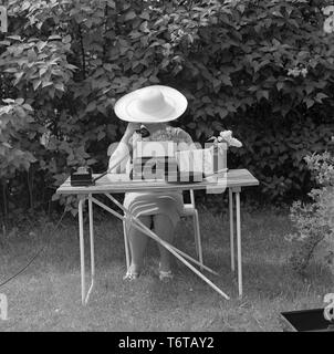 Summer in the 1960s. A lady is sitting in the garden writing on a typewriter. At the same time she is speaking on the phone. A guess is that she is trying to get some work done on her vacation.  Sweden 1961 Stock Photo