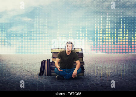 Excited student guy sitting relaxed on the floor listening to music on earphones, leaning on a pile of books, holding phone with calm face with closed Stock Photo