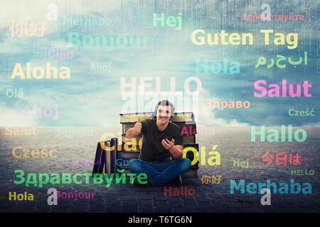 Excited student guy seated near a pile of books, hold his phone, learn different languages on earphones. Thumb up positive feedback, speaks fluently m Stock Photo