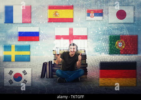 Excited student guy seated near a pile of books, holding phone, learn different languages on headphones. Thumb up positive feedback, speaks fluently m Stock Photo