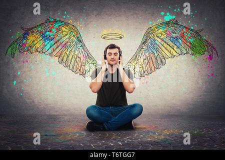 Music gives you colorful wings. Relaxed casual guy seated on the floor listening favourite music on headphones, holding hands to ears dreaming being i Stock Photo
