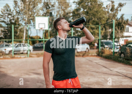 Male athlete, in the summer in the city on the sports ground. In hand a shaker with water and protein. Drinks, resting after a workout fitness in Stock Photo