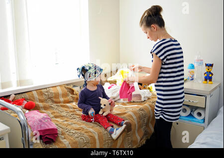 Woman changing clothes to baby girl sitting on a bed of a hospital room. May 18,2018. Kiev,Ukraine Stock Photo
