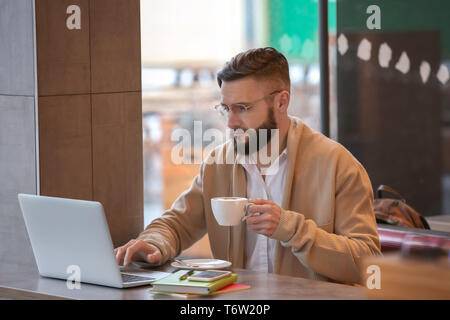 Young freelancer with cup of coffee and laptop working in cafe Stock Photo