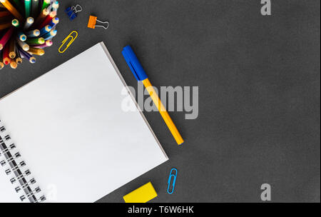 Open white blank spiral paper notebook with pen, colored pencils, eraser, paper clump, paper clips on empty dark background top view Stock Photo