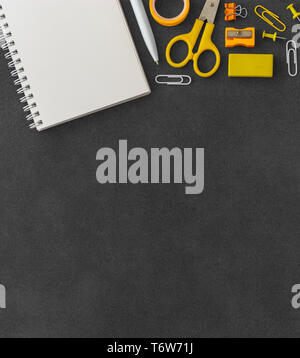 Blank white spiral paper notebook with scissors, paper clump, paper clips, push pins, pen, sharpener and eraser on empty dark background yellow theme Stock Photo