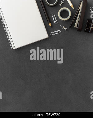Blank white spiral paper notebook with stapler, paper clump, paper clips, push pins and colored pencils on empty dark background top view black theme Stock Photo