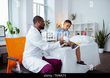 Dark-skinned children doctor looking at leg of little boy Stock Photo