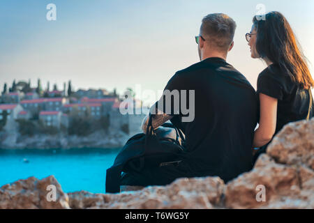 Young couple sitting on cliff above sea and looking at european town on island. Sunset time. Montenegro, Sveti Stefan. Saint Stefan. Travelers and blo Stock Photo