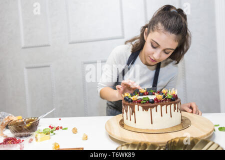 A confectioner stands next to a cooked biscuit cake with white cream, decorated with chocolate and berries. Cake stands on a wooden stand on a white Stock Photo