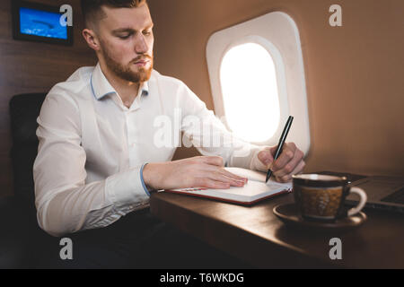 attractive and successful businessman in a white shirtt working sitting in the chair of his private jet. Stock Photo