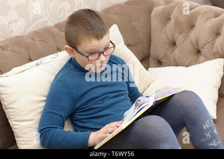 boy in glasses reading a thick book while sitting on the couch Stock Photo