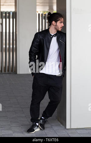 Bearded handsome young man in a black leather jacket with a stylish haircut stands and leans on the wall, looking away. Stock Photo