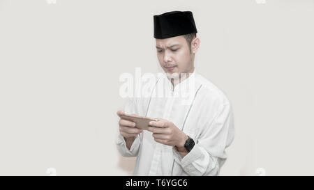 portrait of handsome asian muslim man with head cap and using smartphone and seriously playing video game, muslim on e sport concept - image Stock Photo