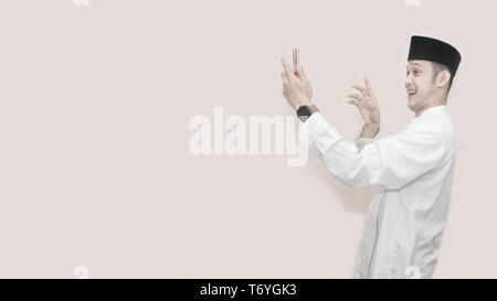 portrait of handsome asian muslim man with head cap and using smartphone taking a video call and waving smile at phone -image Stock Photo