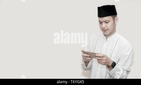portrait of handsome asian muslim man with head cap and using smartphone and seriously playing video game - image Stock Photo