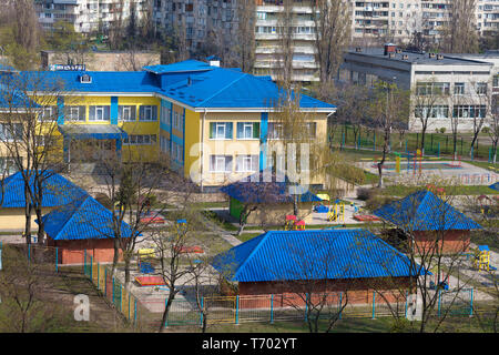 The bright blue roof of the kindergarten is illuminated by the spring sun against the background of gray urban high-rise buildings Stock Photo