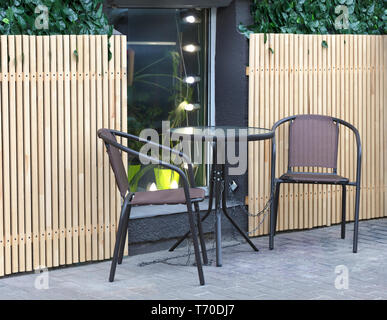 two chairs and a glass round coffee table stand on the street near the window of a cafe on the background of a decorative wooden fence and chained wit Stock Photo