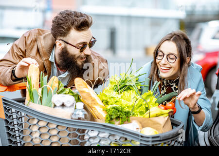 Young and happy couple with shopping cart full of fresh and healthy food outdoors Stock Photo