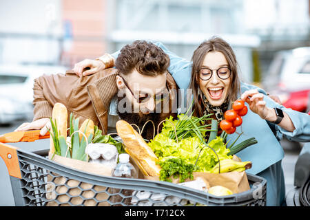 Young and happy couple with shopping cart full of fresh and healthy food outdoors Stock Photo