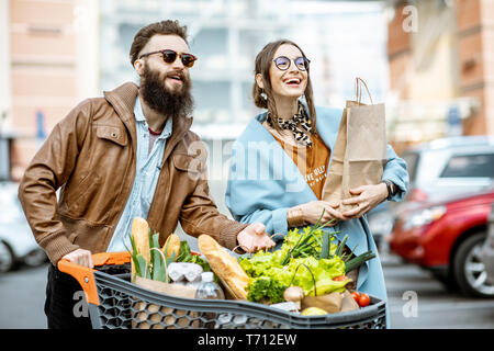 Young and happy couple walking with shoopping cart full of healthy food on the outdoor parking near the supermarket Stock Photo