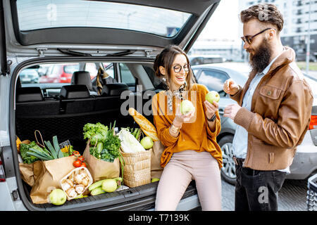 Young stylish couple having fun while sitting on the car trunk full of fresh and healthy food on the supermarket parking outdoors Stock Photo