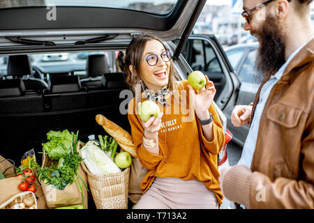 Young stylish couple having fun while sitting on the car trunk full of fresh and healthy food on the supermarket parking outdoors Stock Photo