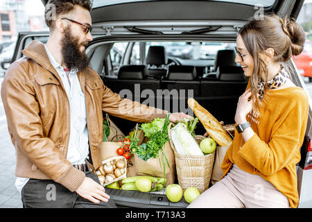 Young stylish couple having fun while sitting on the car trunk full of fresh and healthy food on the supermarket parking outdoors Stock Photo