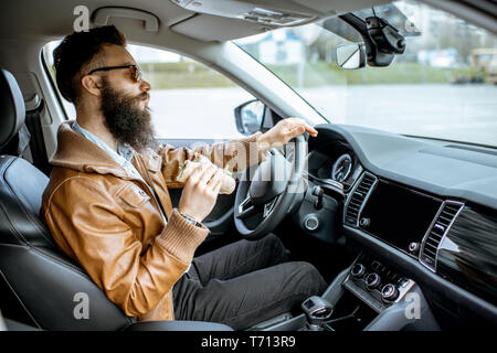 Stylish bearded man having a snack with tasty sandwich while driving a car in the city Stock Photo
