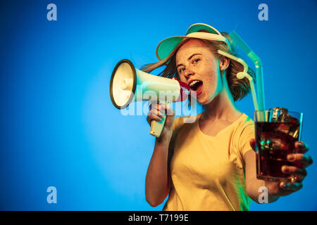 Beautiful female half-length portrait isolated on blue neon lights studio background. Woman in green cap with the drink and mouthpiece. Facial expression, summer, weekend concept. Trendy colors. Stock Photo