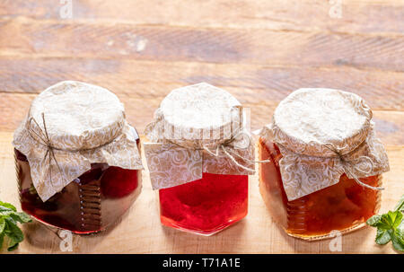 Three small glass jars with homemade cherry, strawberry and apple jam on wooden background with copy space. Jars are closed with homemade paper lids.  Stock Photo