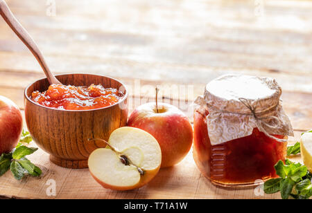 Wooden bowl and small glass jar closed with homemade paper lid with homemade apple jam and apples and green mint on wooden background with copy space. Stock Photo
