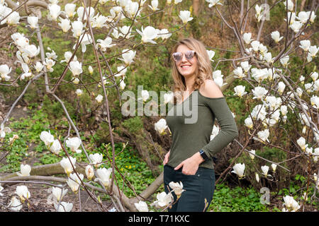 Young blonde woman near blossoming magnolia flowers tree in spring park on sunny day. Magnolia trees. Beautiful happy girl enjoying smell in a Stock Photo