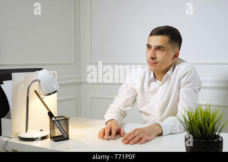 Office worker young man in white shirt is procrastinating sitting at work at the office table. Dreaming about something. Stock Photo