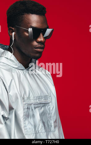 Trendy fashion portrait of man wearing silver outfit and stylish sunglasses. Man in silver hooded shirt wearing earphones and sunglasses against red b Stock Photo