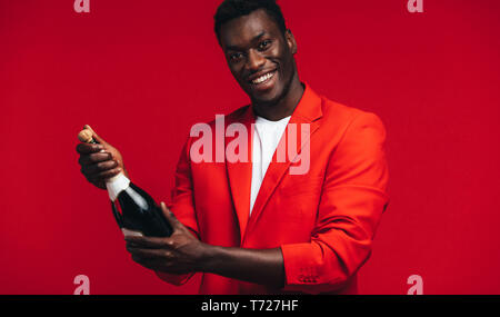 Handsome young african man about to open a champagne bottle against red background. Black guy celebrating with champagne. Stock Photo