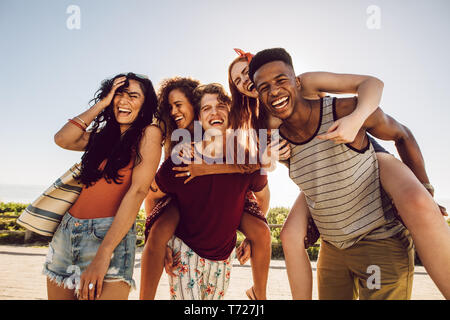 Group of happy friends having fun together outdoors. Men piggybacking female friends and laughing. Summer vacations. Stock Photo