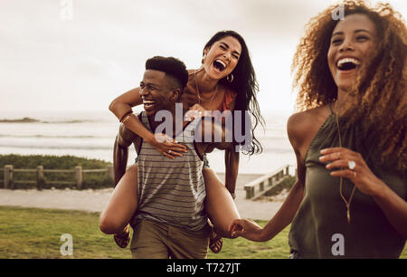Excited young man carrying his girlfriend on his back with female friend walking in front. Diverse group of friends enjoying on a vacation. Stock Photo