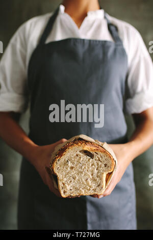 Cropped shot of female baker in uniform holding fresh bread.  Woman baker holding a bread loaf in bakery. Stock Photo