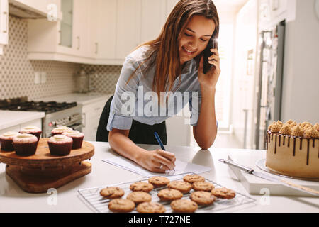Young pastry chef standing at kitchen counter taking order. Happy chef talking on phone and making notes on paper. Stock Photo