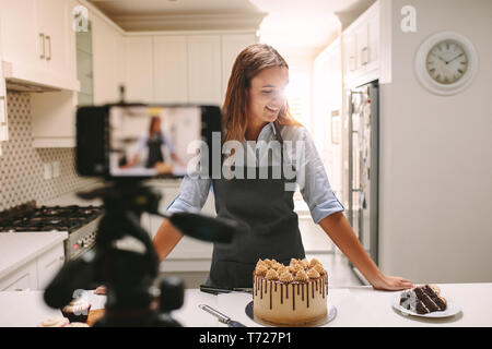 Smiling young woman standing at kitchen counter with pastries recording a content for her food blog channel. Stock Photo