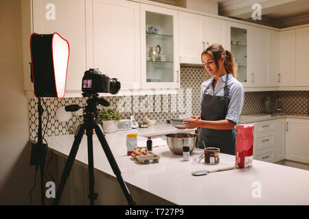 Young woman wearing apron filming herself preparing cake in the kitchen. Pastry chef recording content for the food and baking vlog using a camera mou Stock Photo
