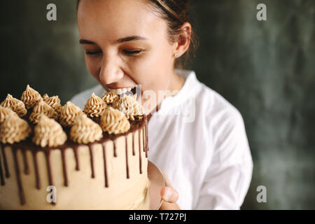 Female confectioner eating whole cake. Woman chef holding a big cake in hand and taking a bite. Stock Photo