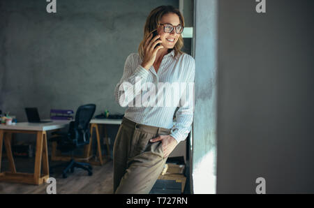 businesswoman talking on cell phone and smiling. Female entrepreneur standing by a window and talking over mobile phone in office Stock Photo