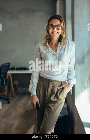 Positive businesswoman standing in office. Woman in casuals standing by window in office looking at camera. Stock Photo