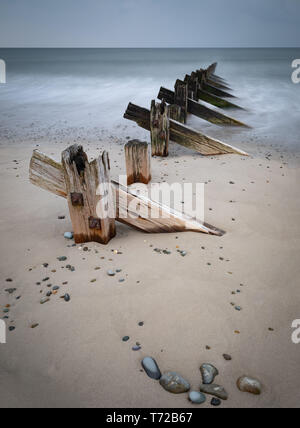 Long exposure image of weathered wooden groyn posts leading from a sandy beach with pebbles into the sea. The waves are smoothed by the long exposure. Stock Photo