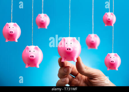 Human's Hand Holding Hanging Pink Piggy Bank Against Blue Background Stock Photo