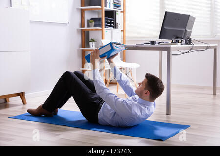 Businessman Doing Situps In Office Holding Folder File In Hand Stock Photo